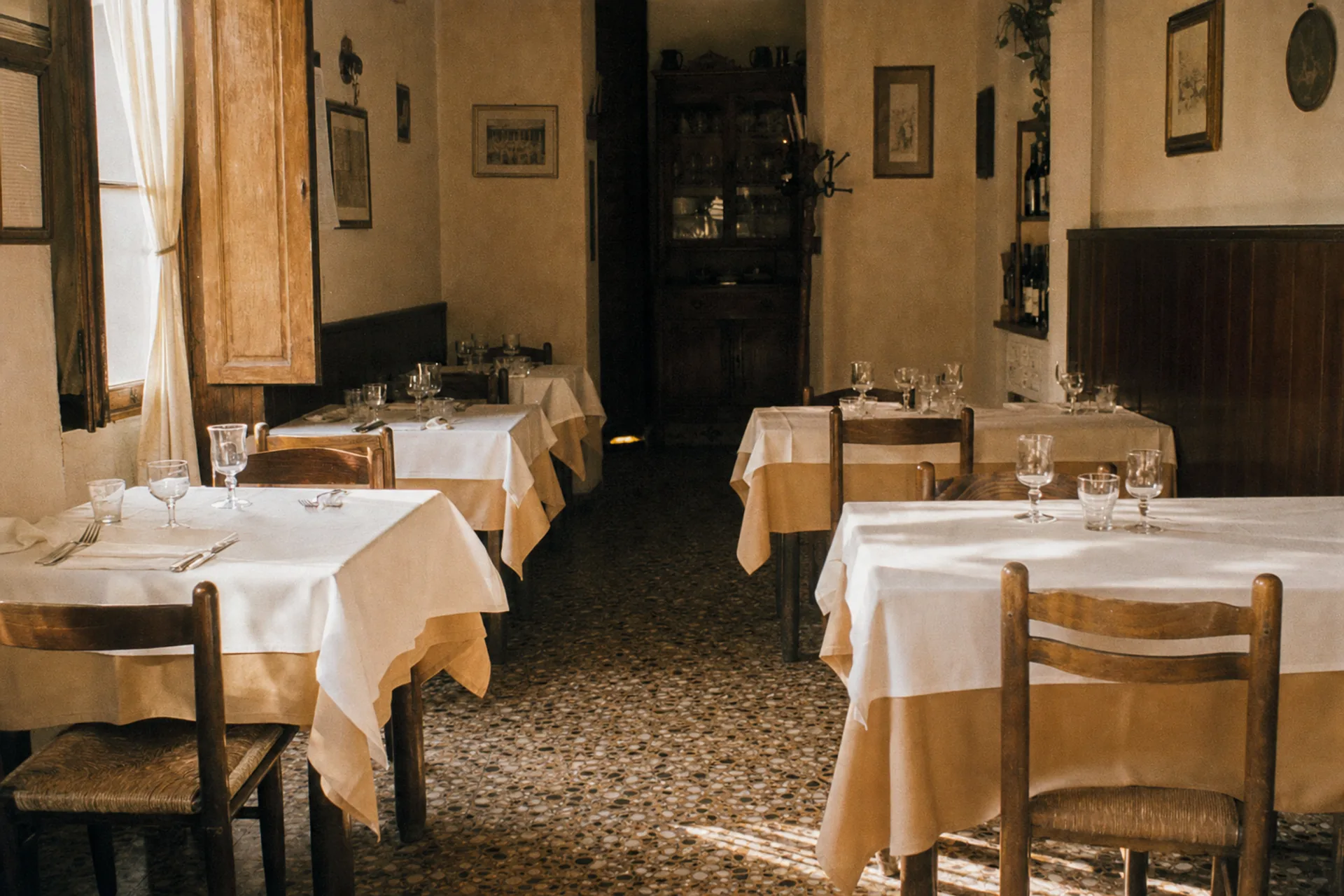 Petite salle de trattoria en fin d'après-midi, tables dressées de nappes blanches, lumière chaude par la fenêtre.